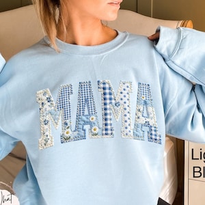May include: Light blue sweatshirt with the word "MAMA" in patchwork letters. The letters feature floral, gingham, and polka dot patterns in shades of blue and white. The sweatshirt is a light blue color.