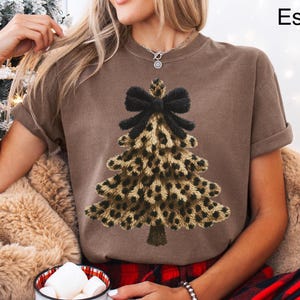 May include: A brown t-shirt with a leopard print Christmas tree design and a black bow. The shirt is part of the "Lovin What U Wear" collection. The model is wearing the shirt while sitting on a fluffy, tan-coloured cushion.