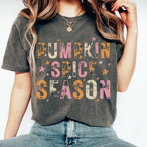 Może przedstawiać: Szary t-shirt z napisem "Pumpkin Spice Season" w kolorze pomarańczowym, r&oacute;żowym i ż&oacute;łtym, z efektem postarzenia. Litery otoczone są małymi złotymi gwiazdkami.