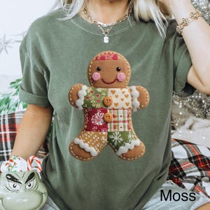 May include: A moss green t-shirt featuring a gingerbread man design. The gingerbread man is made of various fabric patterns and has button details. The text "Moss" is printed on the shirt. The shirt is part of the "Lovin What U Wear" collection.