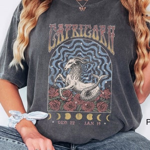 May include: A dark gray t-shirt with a vintage-style graphic of a sea goat with the text "Capricorn" above it. The graphic also includes roses and crescent moons with the dates "Dec 22 - Jan 19".