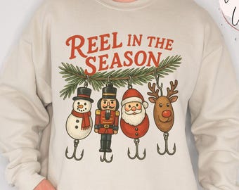 May include: Sand-colored sweatshirt with the text "Reel in the Season" above a row of Christmas-themed fishing lures: a snowman, nutcracker, Santa, and reindeer. The sweatshirt has long sleeves and a crew neck.