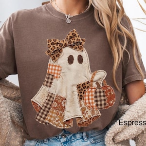 May include: A brown short-sleeve t-shirt featuring a graphic of a ghost wearing a leopard print bow and holding a pumpkin. The ghost is decorated with various patchwork patterns in shades of brown, orange, and cream. The word Espresso is printed.