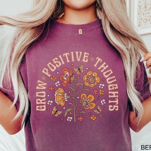 Op de afbeelding: Een paars T-shirt met een grafisch ontwerp van bloemen en vlinders. De tekst "Grow Positive Thoughts" staat in een cirkel rond de bloemen.