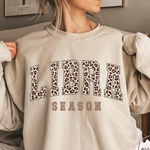 Könnte beinhalten: Ein sandfarbenes Sweatshirt mit Rundhalsausschnitt und dem Text LIBRA SEASON auf der Vorderseite. Das Wort LIBRA ist in großen Blockbuchstaben mit einem braun-schwarzen Leopardenmuster dargestellt, während das Wort SEASON in einfarbigem Braun gehalten ist.