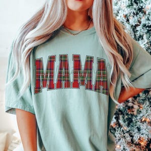 May include: A light green t-shirt with the word "MAMA" in large, red and green plaid letters across the front. The shirt is a relaxed fit and has short sleeves. The text "Bay" is at the bottom right of the image.