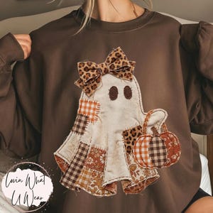 Op de afbeelding: Een donkerchocoladebruine sweater met ronde hals en een centrale applicatie van een spookfiguur. Het spook is versierd met diverse stoffen patches in ruit-, bloemen- en luipaardprints, afgewerkt met een bijpassende strik in luipaardprint.