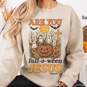 May include: Sand-colored sweatshirt with a Halloween-themed graphic. The design features a pumpkin, ghosts, a cross, and the text "ARE YOU fall-o-ween JESUS Matthew 4:19" in orange and black. The sweatshirt is a long-sleeved crew neck.