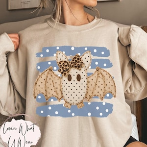 May include: A sand-colored crewneck sweatshirt featuring a central graphic of a beige bat with brown polka dots and a leopard print bow. The bat is positioned over a blue horizontal striped background with white polka dots. The word Sand is visible.
