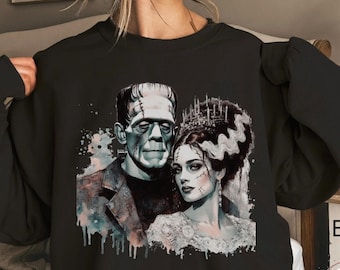 May include: A black long-sleeve sweatshirt featuring a graphic print of Frankenstein's monster and his bride. The design includes watercolor-style paint splatters in muted tones, with the characters depicted in a detailed, artistic illustration style.