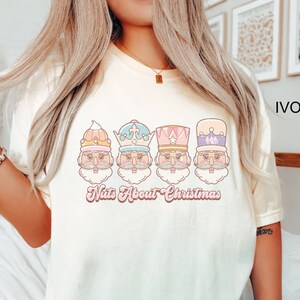 May include: A white t-shirt with a pink, blue, and yellow illustration of four nutcracker faces. The text "Nuts About Christmas" is written in pink below the illustration. The shirt is on a model wearing blue jeans.