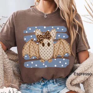 May include: A brown short-sleeve t-shirt featuring a graphic of a tan bat with dark polka dots and a leopard print bow. The bat is centered over a blue rectangular background with white polka dots. The text Espresso is printed on the shirt.
