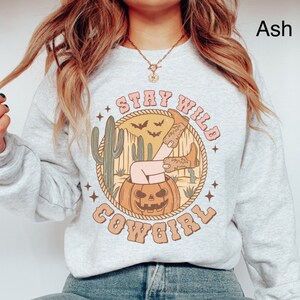 May include: A light grey sweatshirt with a graphic print of a woman's legs sticking out of a jack-o-lantern. The graphic also includes a cactus, bats, and the text "Stay Wild Cowgirl".