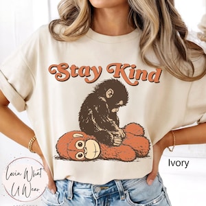 Pode incluir: Camiseta cor marfim com a frase "Stay Kind" em escrita laranja. A camiseta apresenta uma ilustração marrom de um macaco abraçando um urso de pelúcia. A camiseta tem um corte folgado.