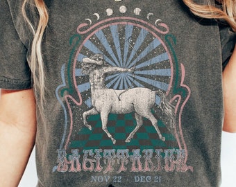 May include: A dark gray t-shirt with a vintage-style graphic of a Sagittarius zodiac sign. The graphic features a centaur with an arrow and bow, surrounded by stars and a sunburst. The text "Sagittarius" and "Nov 22 - Dec 21" is printed below the graphic.
