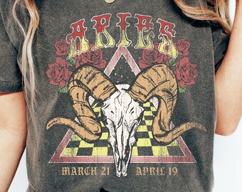May include: Dark gray Aries graphic t-shirt. The design features a ram skull, roses, and the word "ARIES" in a vintage style. The shirt also includes the dates "MARCH 21 - APRIL 19".