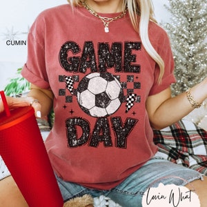 May include: A cumin-colored t-shirt with the words "GAME DAY" in a distressed black font, featuring a soccer ball graphic. The shirt also includes checkered flag accents, perfect for sports fans.