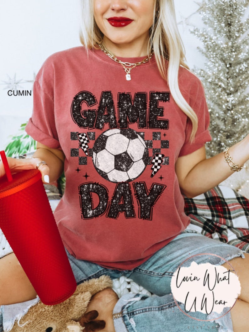 May include: A cumin-colored t-shirt with the words "GAME DAY" in a distressed black font, featuring a soccer ball graphic. The shirt also includes checkered flag accents, perfect for sports fans.