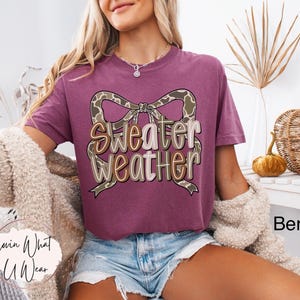 May include: Berry-colored t-shirt with the text "Sweater Weather" in a decorative font, featuring a bow design. The shirt is part of a fall-themed collection. The image also includes a decorative pumpkin and a woven basket.
