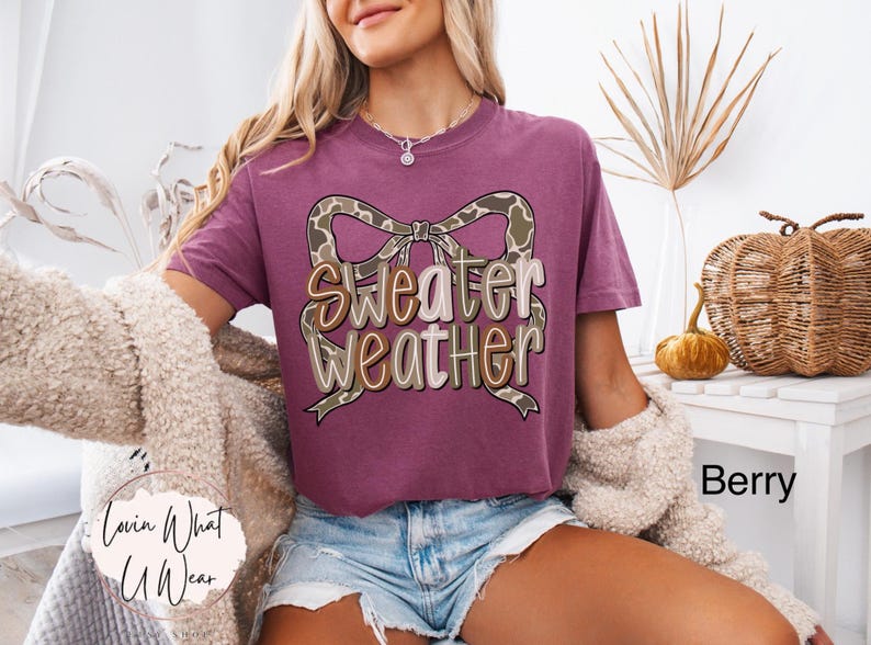 May include: Berry-colored t-shirt with the text "Sweater Weather" in a decorative font, featuring a bow design. The shirt is part of a fall-themed collection. The image also includes a decorative pumpkin and a woven basket.