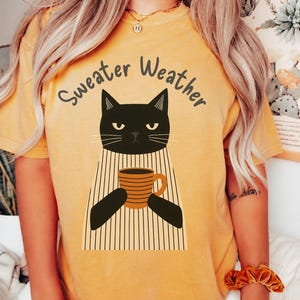 May include: Mustard-colored t-shirt with a graphic of a black cat holding a coffee mug. The text "Sweater Weather" is arched above the cat. The shirt has a relaxed fit and short sleeves, perfect for fall.