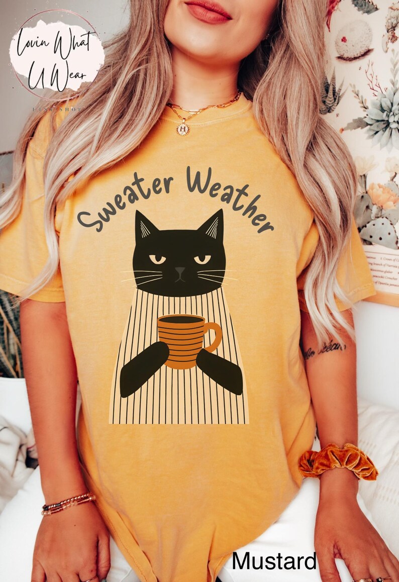May include: Mustard-colored t-shirt with a graphic of a black cat holding a coffee mug. The text "Sweater Weather" is arched above the cat. The shirt has a relaxed fit and short sleeves, perfect for fall.