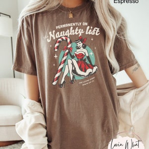 May include: A brown t-shirt with a vintage-style graphic of a woman in a red dress holding a candy cane. The text on the shirt reads "Permanently on the Naughty List" and "And loving every minute of it".