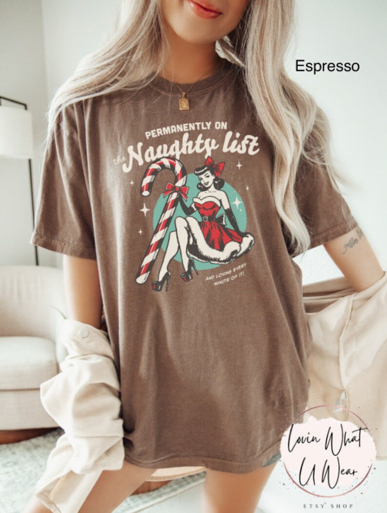 May include: A brown t-shirt with a vintage-style graphic of a woman in a red dress holding a candy cane. The text on the shirt reads "Permanently on the Naughty List" and "And loving every minute of it".