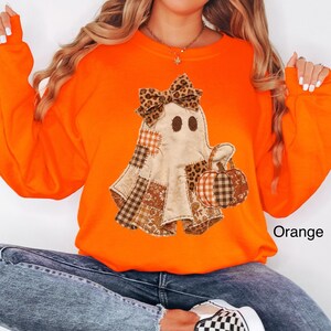 Op de afbeelding: Een feloranje sweatshirt met lange mouwen en een centrale afbeelding van een geest met een strik met luipaardprint. De geest is versierd met diverse bruine en oranje ruitjes- en bloemenstoffen lapjes en houdt een pompoen vast.