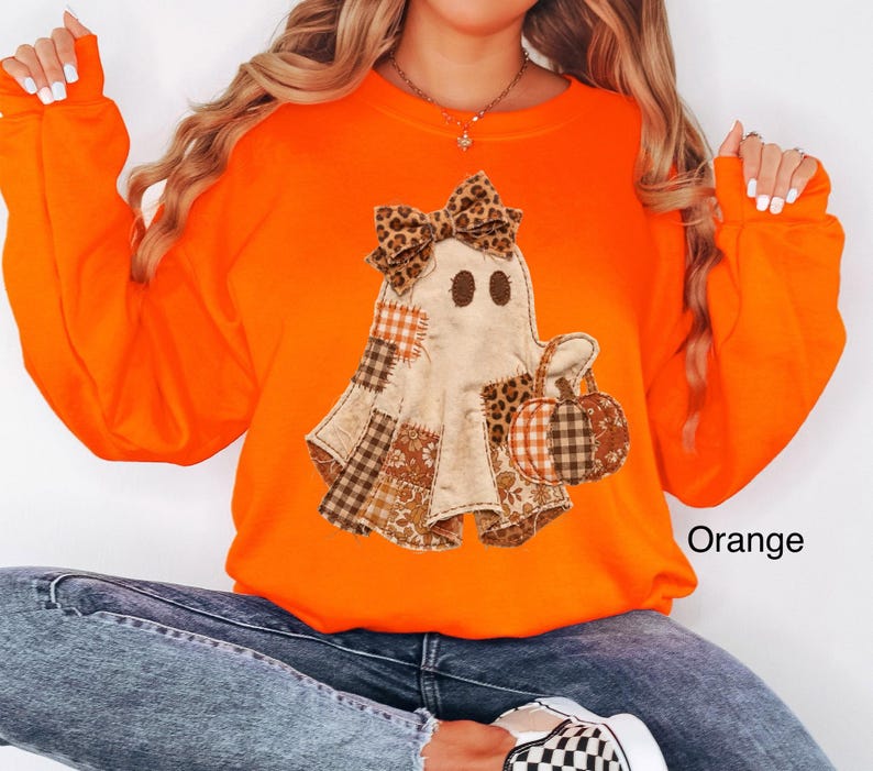 Op de afbeelding: Een feloranje sweatshirt met lange mouwen en een centrale afbeelding van een geest met een strik met luipaardprint. De geest is versierd met diverse bruine en oranje ruitjes- en bloemenstoffen lapjes en houdt een pompoen vast.