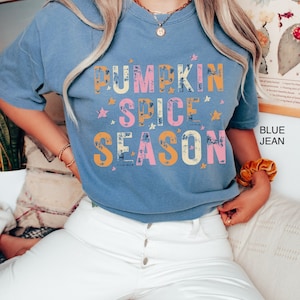 Może przedstawiać: Niebieska koszulka z napisem "Pumpkin Spice Season" w stylu vintage, w kolorze ż&oacute;łtym, r&oacute;żowym i pomarańczowym. Tekst otoczony jest małymi białymi gwiazdkami. Modelka ma na sobie białe jeansy.