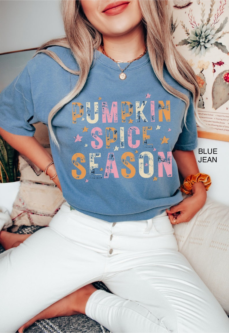 Może przedstawiać: Niebieska koszulka z napisem "Pumpkin Spice Season" w stylu vintage, w kolorze ż&oacute;łtym, r&oacute;żowym i pomarańczowym. Tekst otoczony jest małymi białymi gwiazdkami. Modelka ma na sobie białe jeansy.