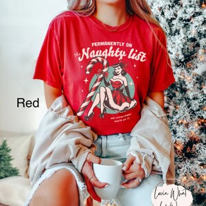 May include: Red t-shirt with a vintage-style graphic of a woman in a red dress holding a candy cane. The text on the shirt reads "Permanently on the Naughty List" and "And loving every minute of it!"
