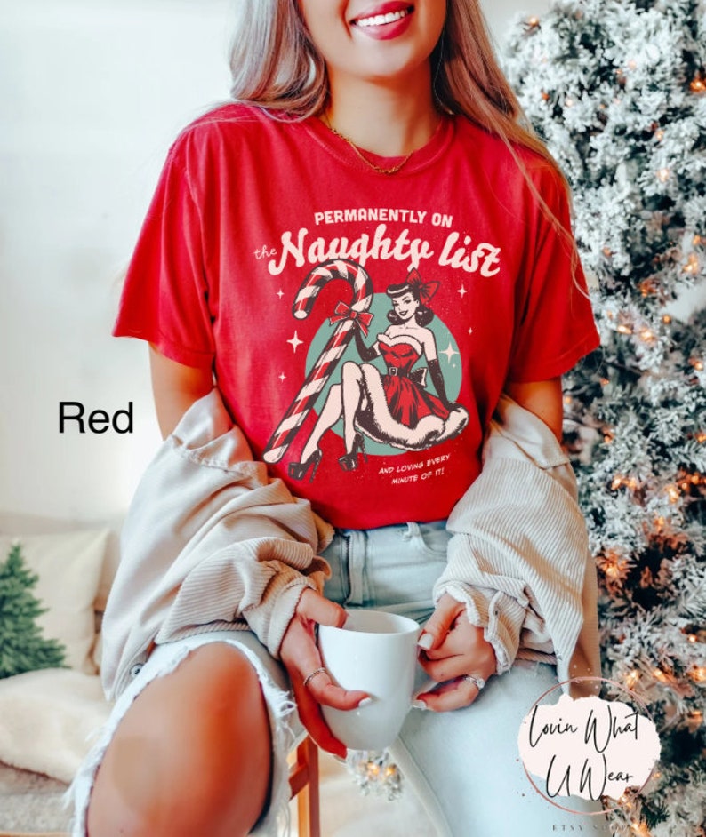 May include: Red t-shirt with a vintage-style graphic of a woman in a red dress holding a candy cane. The text on the shirt reads "Permanently on the Naughty List" and "And loving every minute of it!"