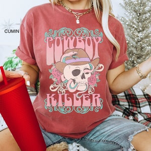 Op de afbeelding: Een rood T-shirt met een vintage-stijl grafische afbeelding van een schedel die een cowboyhoed en een slang draagt. De afbeelding zegt "COWBOY KILLER" in roze en turquoise letters.