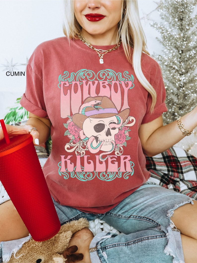 Op de afbeelding: Een rood T-shirt met een vintage-stijl grafische afbeelding van een schedel die een cowboyhoed en een slang draagt. De afbeelding zegt "COWBOY KILLER" in roze en turquoise letters.