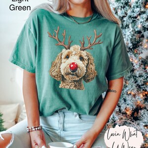 K&ouml;nnte beinhalten: Ein hellgr&uuml;nes T-Shirt mit einem Aufdruck eines braunen und wei&szlig;en Hundes, der eine rote Rentiernase und Geweih tr&auml;gt. Der Text "Lovin What U Wear" ist in der rechten unteren Ecke des Bildes sichtbar.