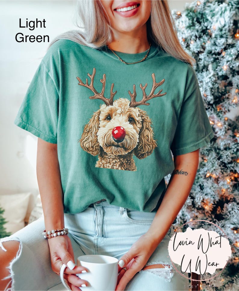 K&ouml;nnte beinhalten: Ein hellgr&uuml;nes T-Shirt mit einem Aufdruck eines braunen und wei&szlig;en Hundes, der eine rote Rentiernase und Geweih tr&auml;gt. Der Text "Lovin What U Wear" ist in der rechten unteren Ecke des Bildes sichtbar.