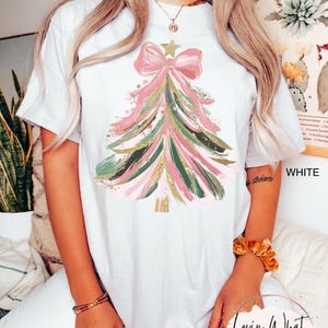 Op de afbeelding: Wit T-shirt met een kerstboomontwerp. De boom is gemaakt van roze, groene en gouden penseelstreken, bekroond met een roze strik en een ster. De tekst "WHITE" is zichtbaar aan de rechterkant van het shirt.