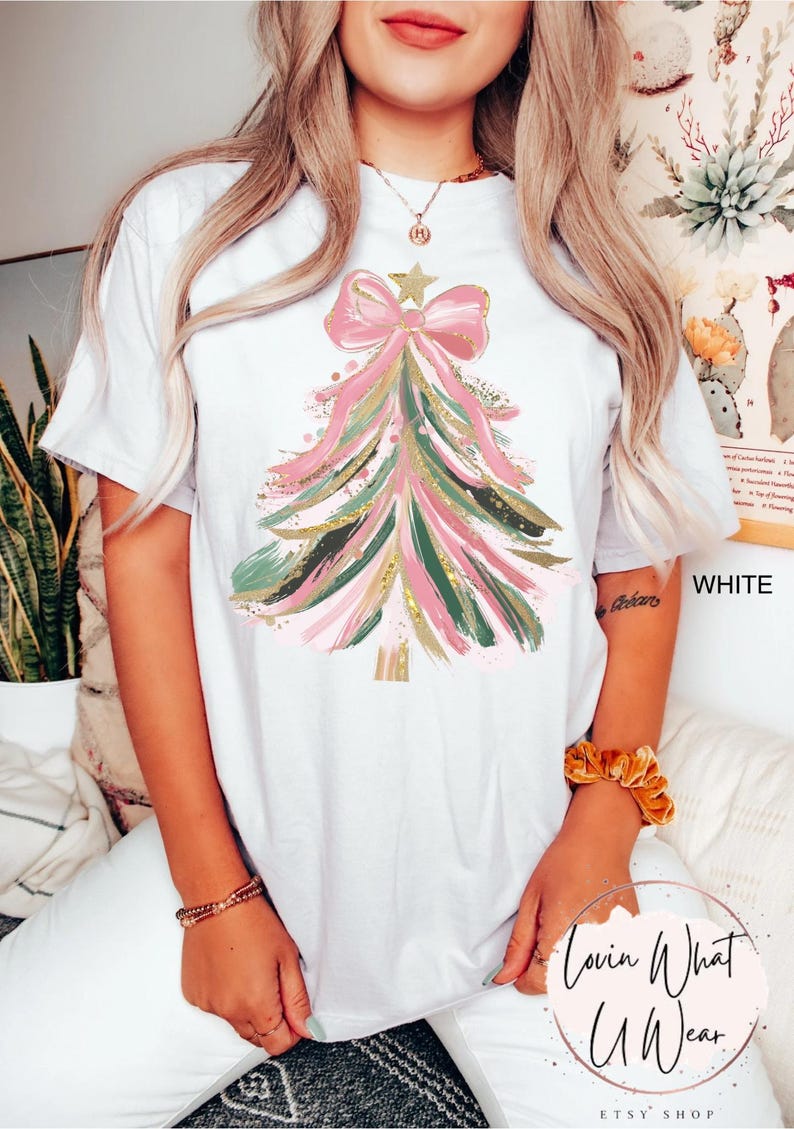 Op de afbeelding: Wit T-shirt met een kerstboomontwerp. De boom is gemaakt van roze, groene en gouden penseelstreken, bekroond met een roze strik en een ster. De tekst "WHITE" is zichtbaar aan de rechterkant van het shirt.