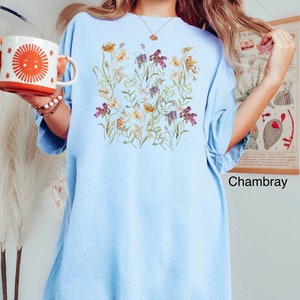 May include: A light blue oversized t-shirt with a floral print featuring a variety of flowers in shades of purple, yellow, and white. The text " Chambray" is printed in black at the bottom right corner of the shirt.