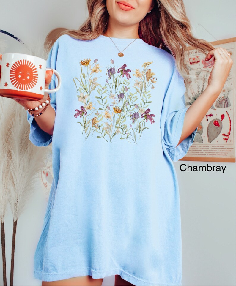 May include: A light blue oversized t-shirt with a floral print featuring a variety of flowers in shades of purple, yellow, and white. The text " Chambray" is printed in black at the bottom right corner of the shirt.