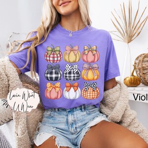 Peut inclure: T-shirt violet avec une grille de neuf citrouilles orn&eacute;es de n&oelig;uds vari&eacute;s. Les citrouilles sont orange, rose et jaune, avec des n&oelig;uds &agrave; carreaux, &agrave; pois et unis. Le texte "Lovin What U Wear" est visible.