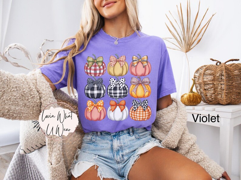 Peut inclure: T-shirt violet avec une grille de neuf citrouilles orn&eacute;es de n&oelig;uds vari&eacute;s. Les citrouilles sont orange, rose et jaune, avec des n&oelig;uds &agrave; carreaux, &agrave; pois et unis. Le texte "Lovin What U Wear" est visible.