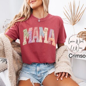 Puede incluir: Camiseta de color carmes&iacute; con la palabra "MAMA" en letras de patchwork. Las letras est&aacute;n hechas de varias telas florales y estampadas. La camiseta tiene mangas cortas y cuello redondo. El texto "Lovin What U Wear" est&aacute; en un c&iacute;rculo a la derecha.