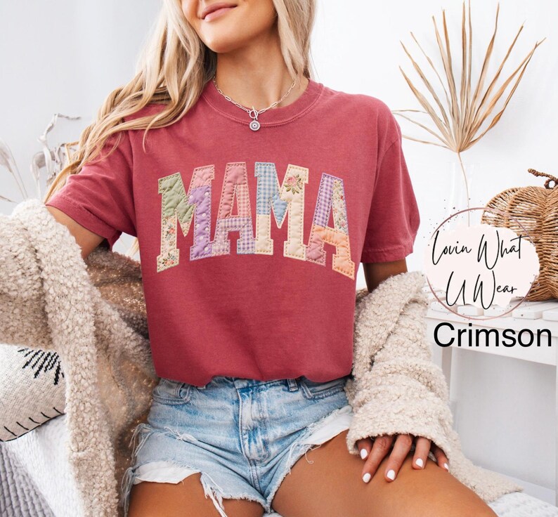 Puede incluir: Camiseta de color carmes&iacute; con la palabra "MAMA" en letras de patchwork. Las letras est&aacute;n hechas de varias telas florales y estampadas. La camiseta tiene mangas cortas y cuello redondo. El texto "Lovin What U Wear" est&aacute; en un c&iacute;rculo a la derecha.