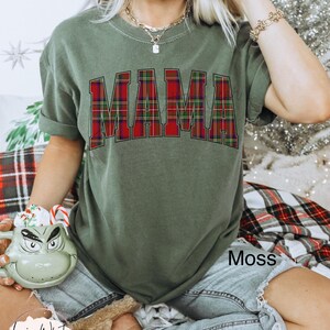 May include: A moss green t-shirt with the word "MAMA" in a red and green plaid design. The shirt is paired with denim shorts. The image also includes a Grinch-themed mug and a plush toy.