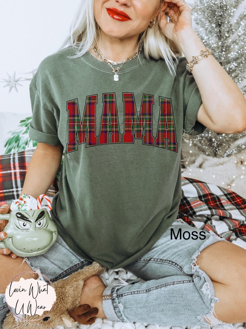 May include: A moss green t-shirt with the word "MAMA" in a red and green plaid design. The shirt is paired with denim shorts. The image also includes a Grinch-themed mug and a plush toy.