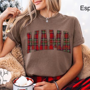 May include: Espresso-colored t-shirt with the word "MAMA" in a red and green plaid design. The shirt has rolled-up sleeves and is part of a holiday-themed collection. The model is wearing a red and black plaid pajama set.