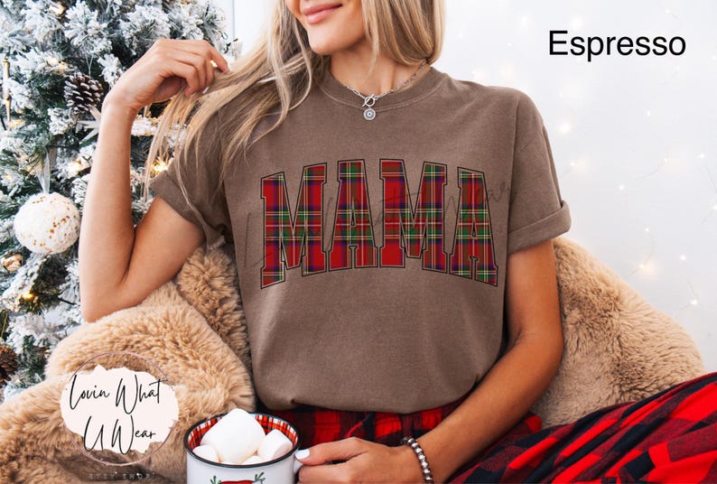 May include: Espresso-colored t-shirt with the word "MAMA" in a red and green plaid design. The shirt has rolled-up sleeves and is part of a holiday-themed collection. The model is wearing a red and black plaid pajama set.